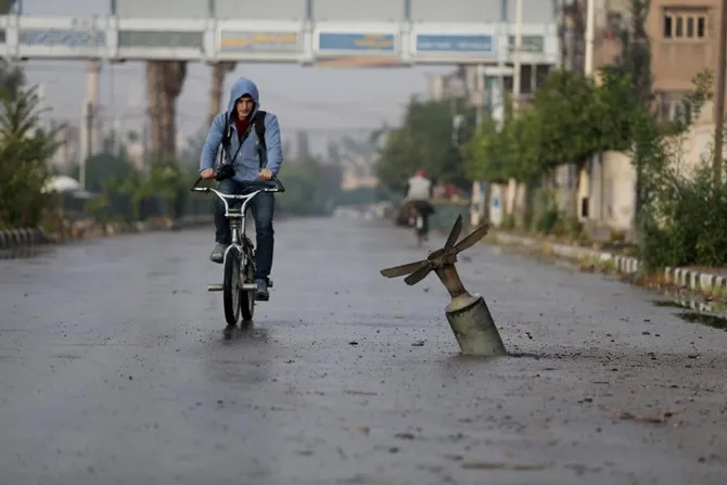 Người dân đạp xe qua đoạn đường có quả bom chưa nổ ở thị trấn Douma, miền Đông Ghouta, Damascus, Syria